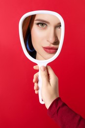 Image of Beautiful woman looking in handheld mirror on red background, closeup