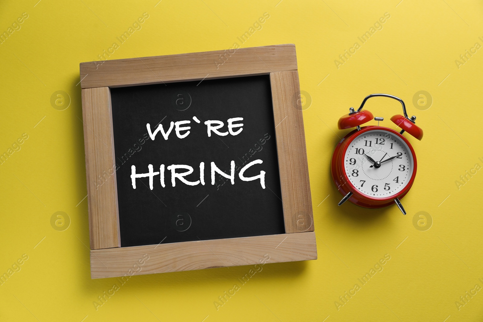 Image of Small chalkboard text We're Hiring and alarm clock on yellow background, top view. Human resource