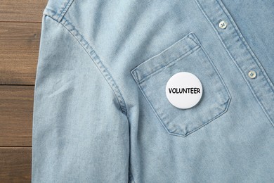 Image of Button badge with word Volunteer attached to denim shirt on wooden background, top view