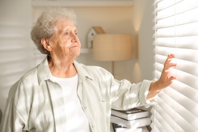 Photo of Loneliness concept. Sad senior woman looking through window blinds at home