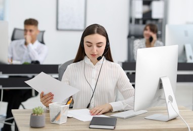 Photo of Saleswoman checking documents while talking to client via headset at desk in office