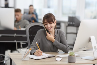 Photo of Saleswoman making notes while talking on phone at desk in office