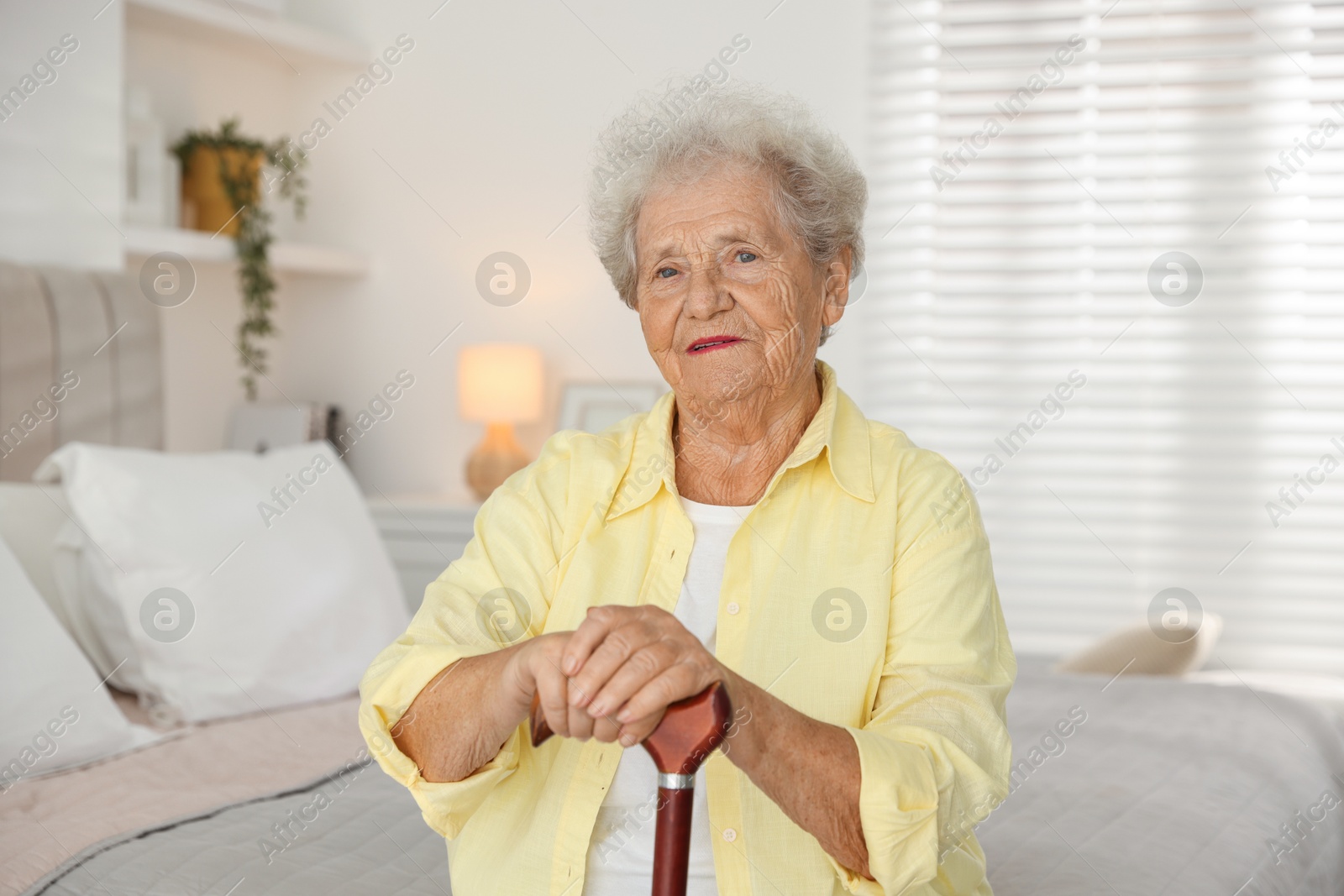 Photo of Senior woman with walking cane sitting on bed at home