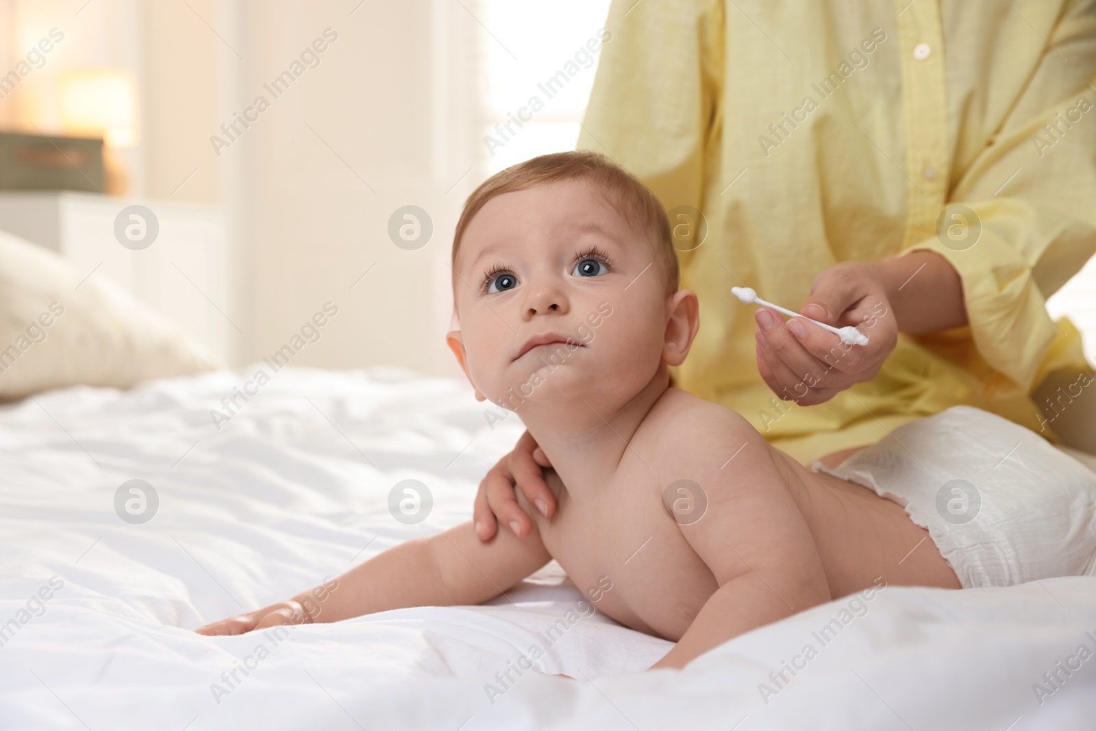 Photo of Mother cleaning ear of her cute little baby with cotton swab on bed, closeup