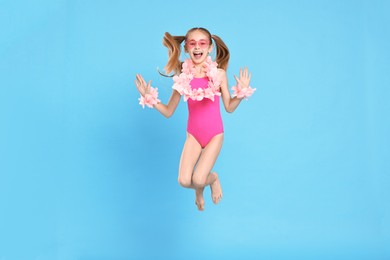 Photo of Happy little girl in beachwear and wreaths of flowers jumping on light blue background