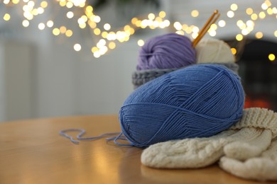 Photo of Colorful yarns, knitted mittens and crochet hooks on wooden table against blurred lights, closeup. Space for text