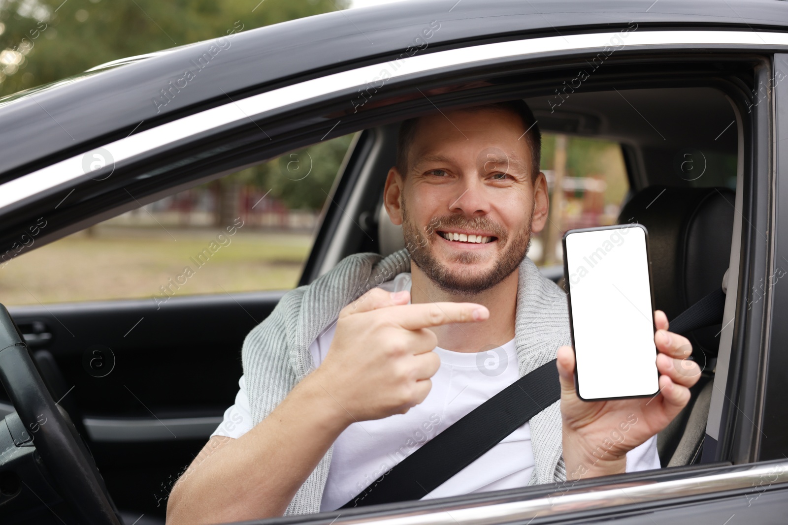 Photo of Happy man pointing at smartphone in modern car, view from outside