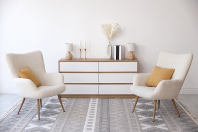 Photo of Soft armchairs and chest of drawers near white wall indoors