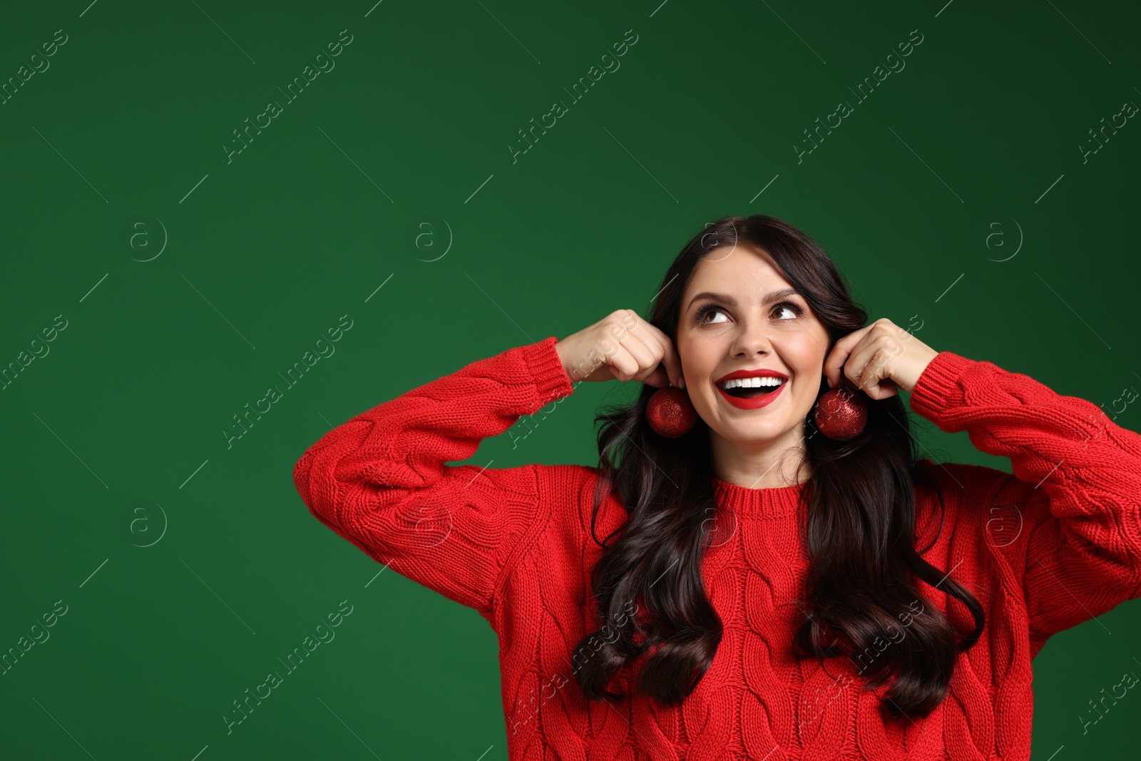 Photo of Happy woman trying Christmas balls as earrings on green background, space for text