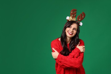 Photo of Happy woman in sweater and reindeer headband on green background, space for text. Christmas celebration