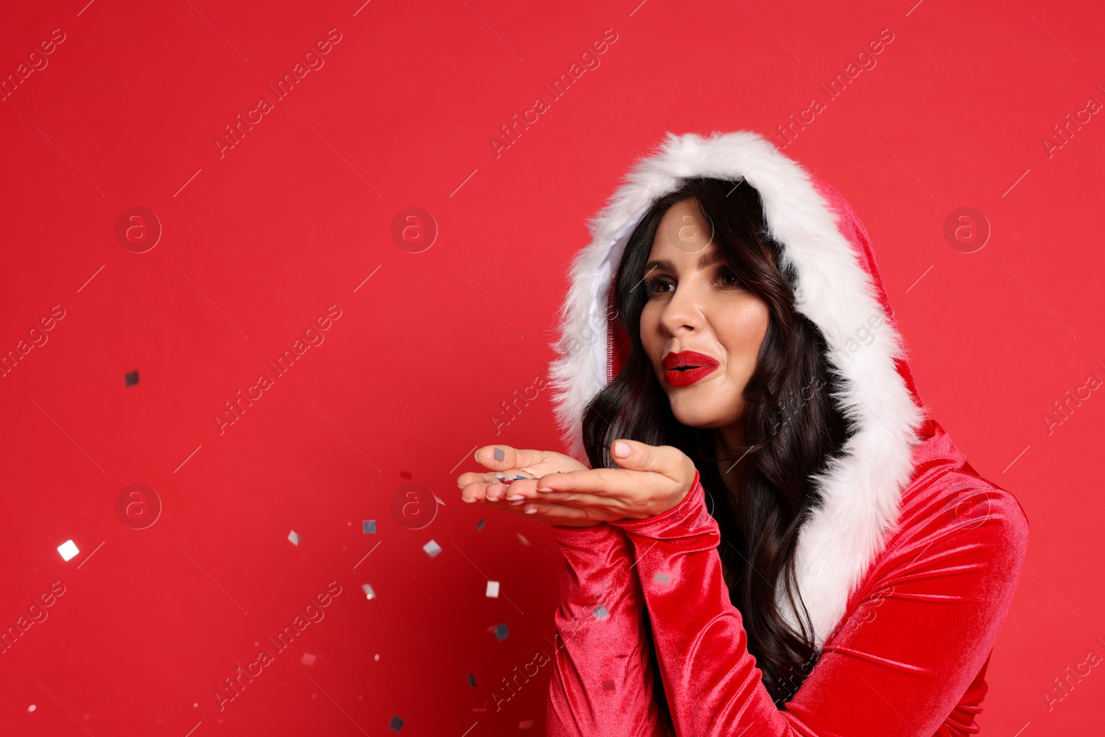 Photo of Beautiful woman in Mrs Claus costume blowing confetti off hands on red background, space for text. Christmas celebration