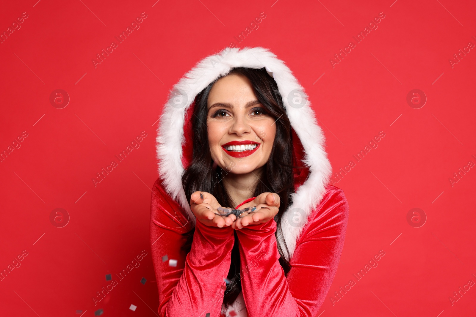 Photo of Beautiful woman in Mrs Claus costume blowing confetti off hands on red background. Christmas celebration