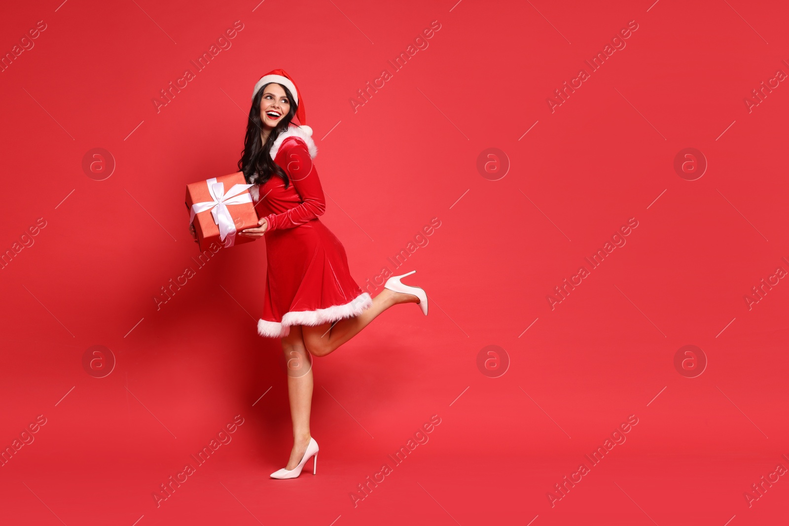 Photo of Beautiful woman in Mrs Claus costume with Christmas gift on red background, space for text