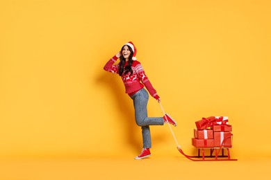 Photo of Woman in Santa hat with Christmas gifts on sledge against yellow background