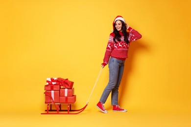 Photo of Woman in Santa hat with Christmas gifts on sledge against yellow background, space for text