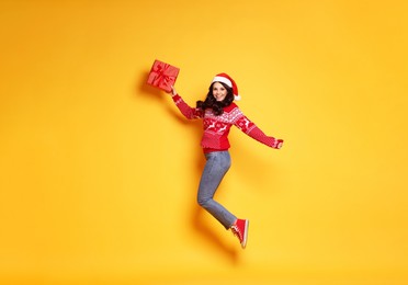 Photo of Woman in Santa hat with Christmas gift jumping on yellow background