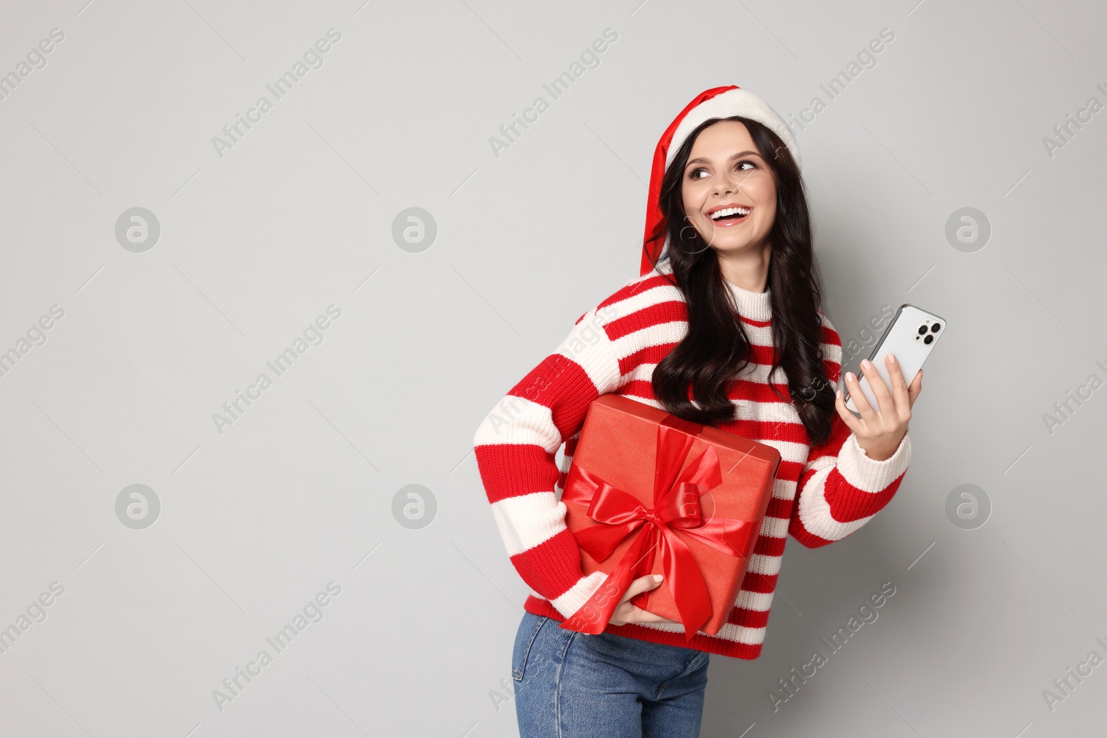 Photo of Woman in Santa hat with Christmas gift and smartphone on light grey background, space for text