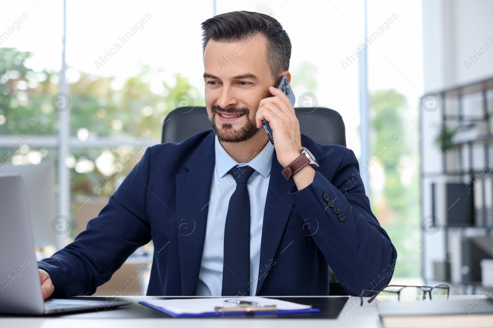 Photo of Happy banker talking on smartphone at table in office