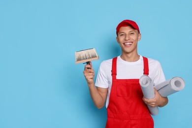 Photo of Young decorator with brush and rolls of wallpaper on light blue background, space for text