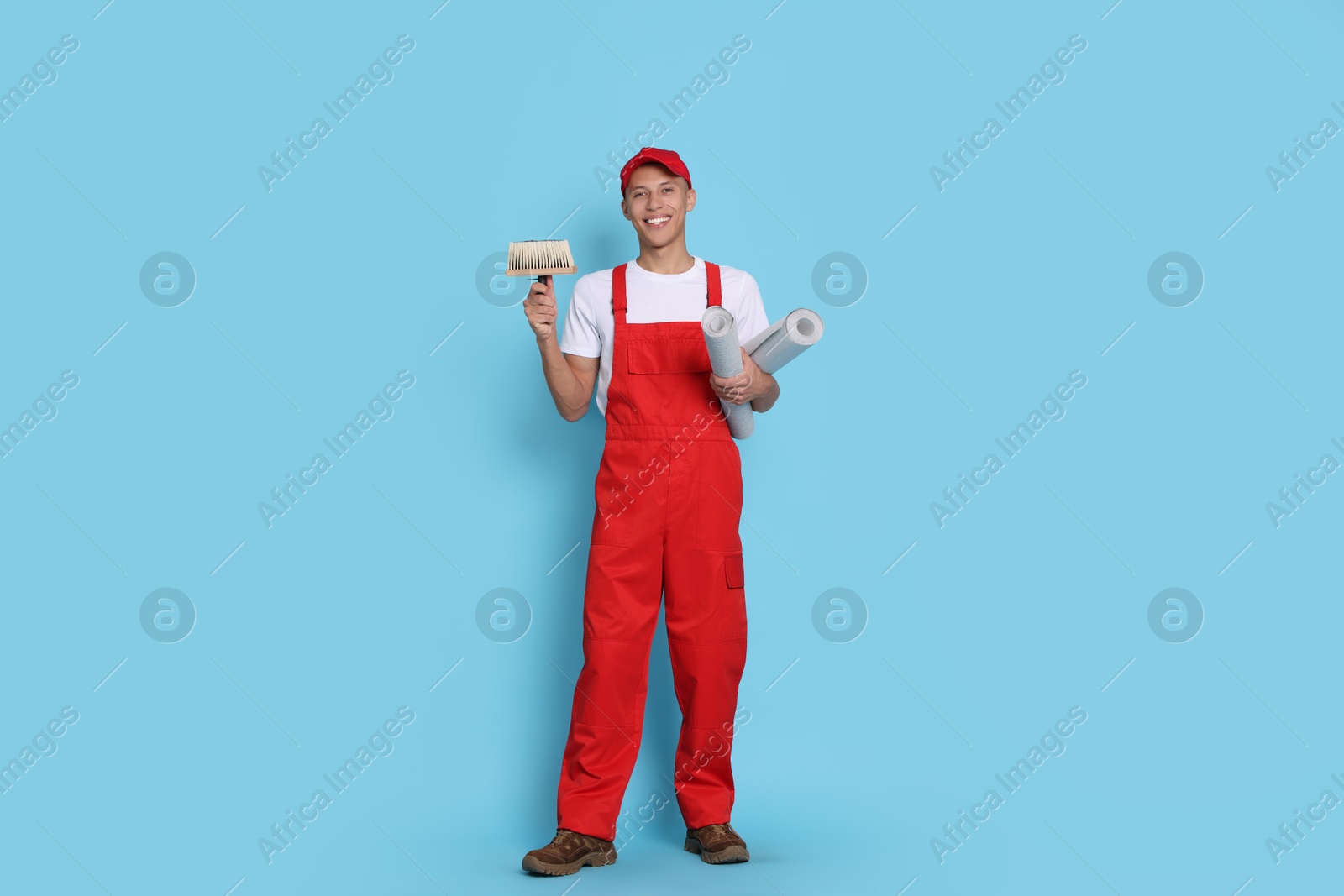 Photo of Young decorator with brush and rolls of wallpaper on light blue background