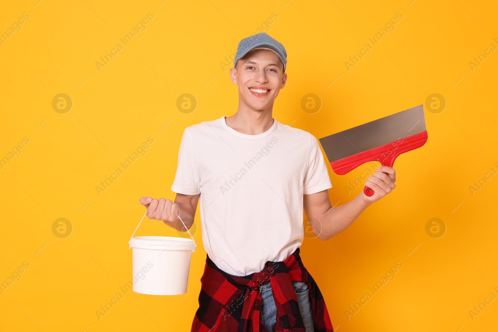 Photo of Portrait of young decorator with putty knife and bucket on orange background