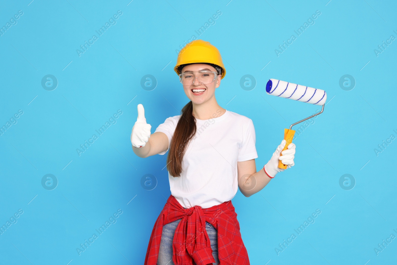 Photo of Portrait of young decorator with paint roller showing thumbs up on light blue background