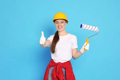 Photo of Portrait of young decorator with paint roller showing thumbs up on light blue background