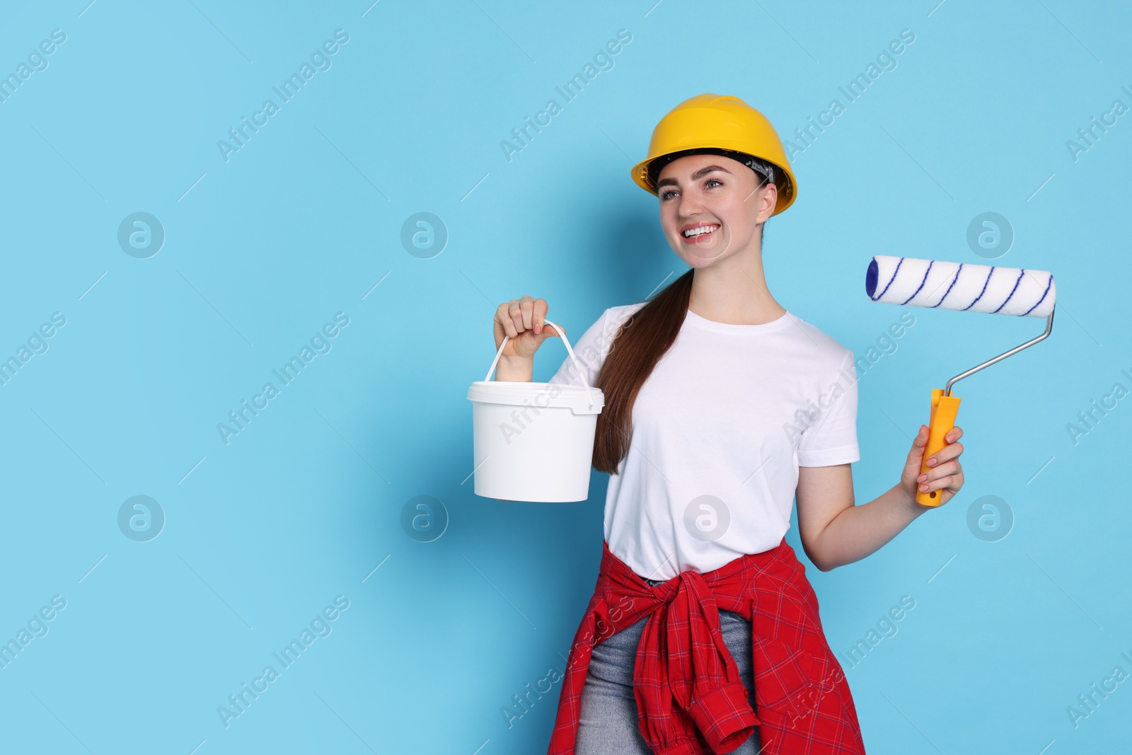 Photo of Portrait of young decorator with paint roller and bucket on light blue background, space for text