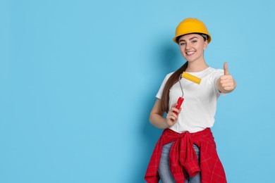 Photo of Portrait of young decorator with paint roller showing thumbs up on light blue background, space for text