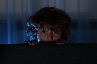 Photo of Fearful boy hiding behind sofa at night