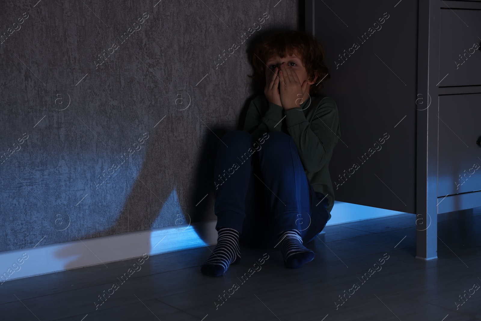 Photo of Scared boy hiding behind chest of drawers at night