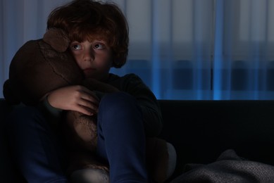 Photo of Scared boy with teddy bear on sofa at night. Space for text