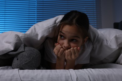 Photo of Fearful girl with toy bunny under duvet in bed at night