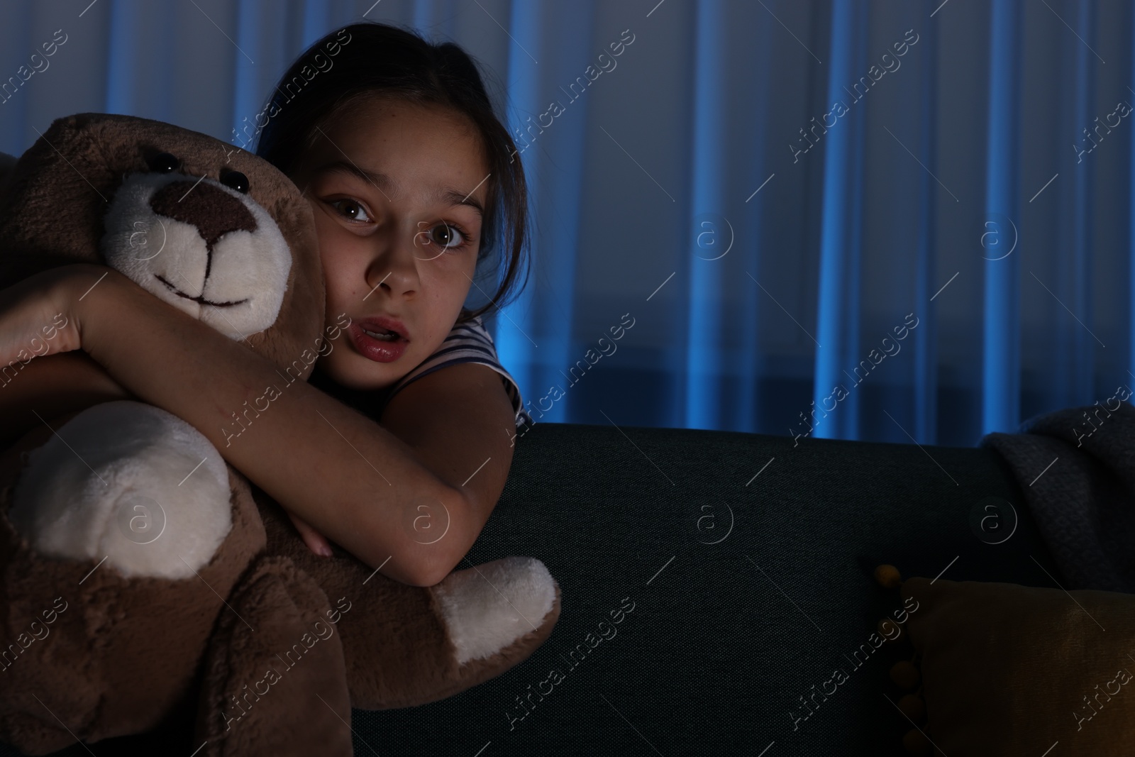 Photo of Scared girl with teddy bear hiding behind sofa at night. Space for text