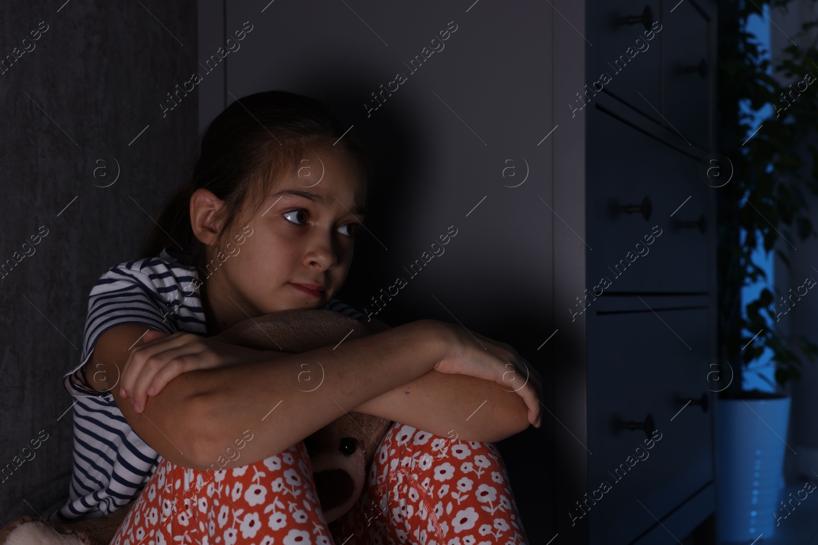 Photo of Scared girl with teddy bear hiding behind chest of drawers in her room at night