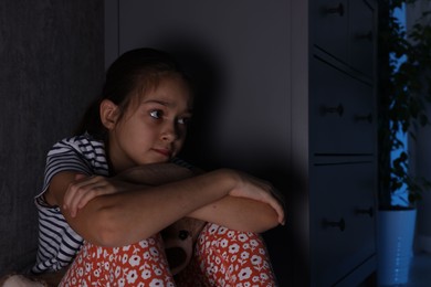 Photo of Scared girl with teddy bear hiding behind chest of drawers in her room at night