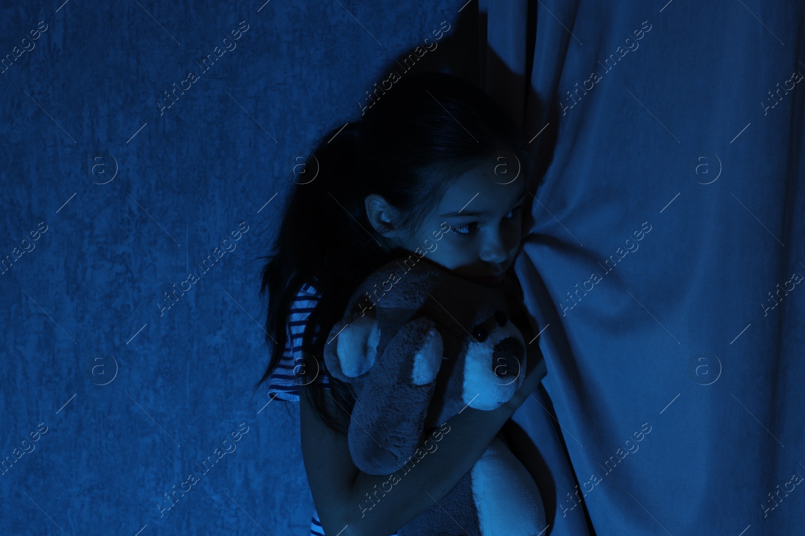 Photo of Scared girl hiding with teddy bear behind curtain at night. Space for text