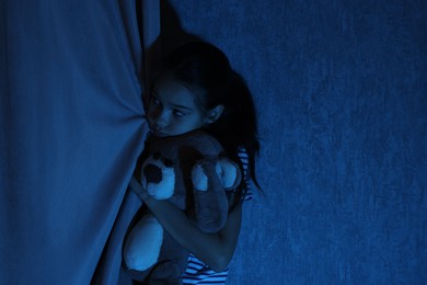 Photo of Scared girl hiding with teddy bear behind curtain at night. Space for text