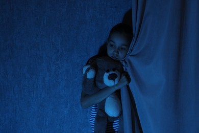 Photo of Scared girl hiding with teddy bear behind curtain at night. Space for text