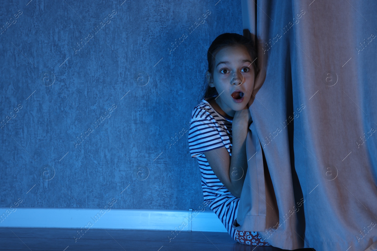 Photo of Scared girl hiding behind curtain at night. Space for text