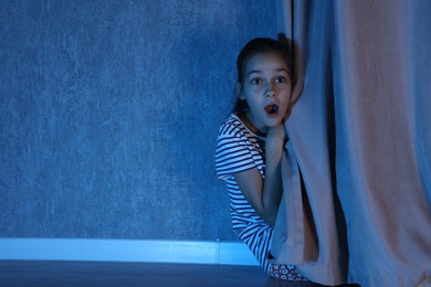 Photo of Scared girl hiding behind curtain at night. Space for text