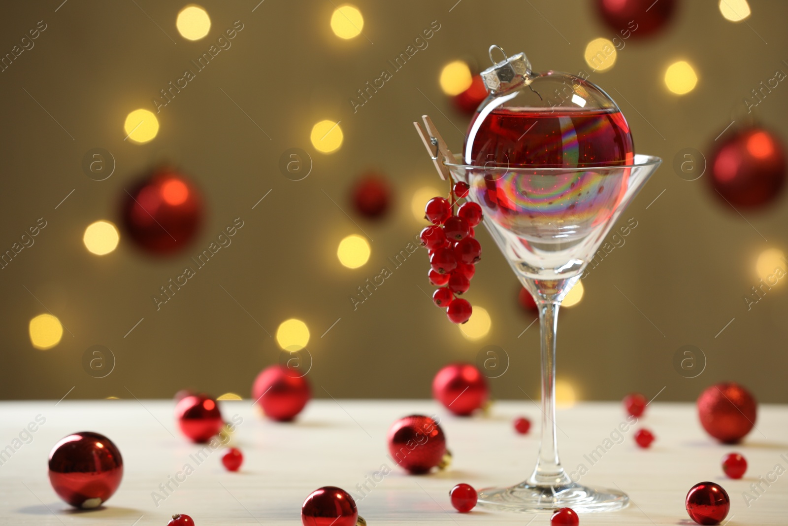 Photo of Creative presentation of Christmas cocktail in bauble and glass on white table against dark beige background with blurred lights. Space for text