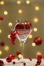 Photo of Creative presentation of Christmas cocktail in bauble and glass on white table against dark beige background with blurred lights