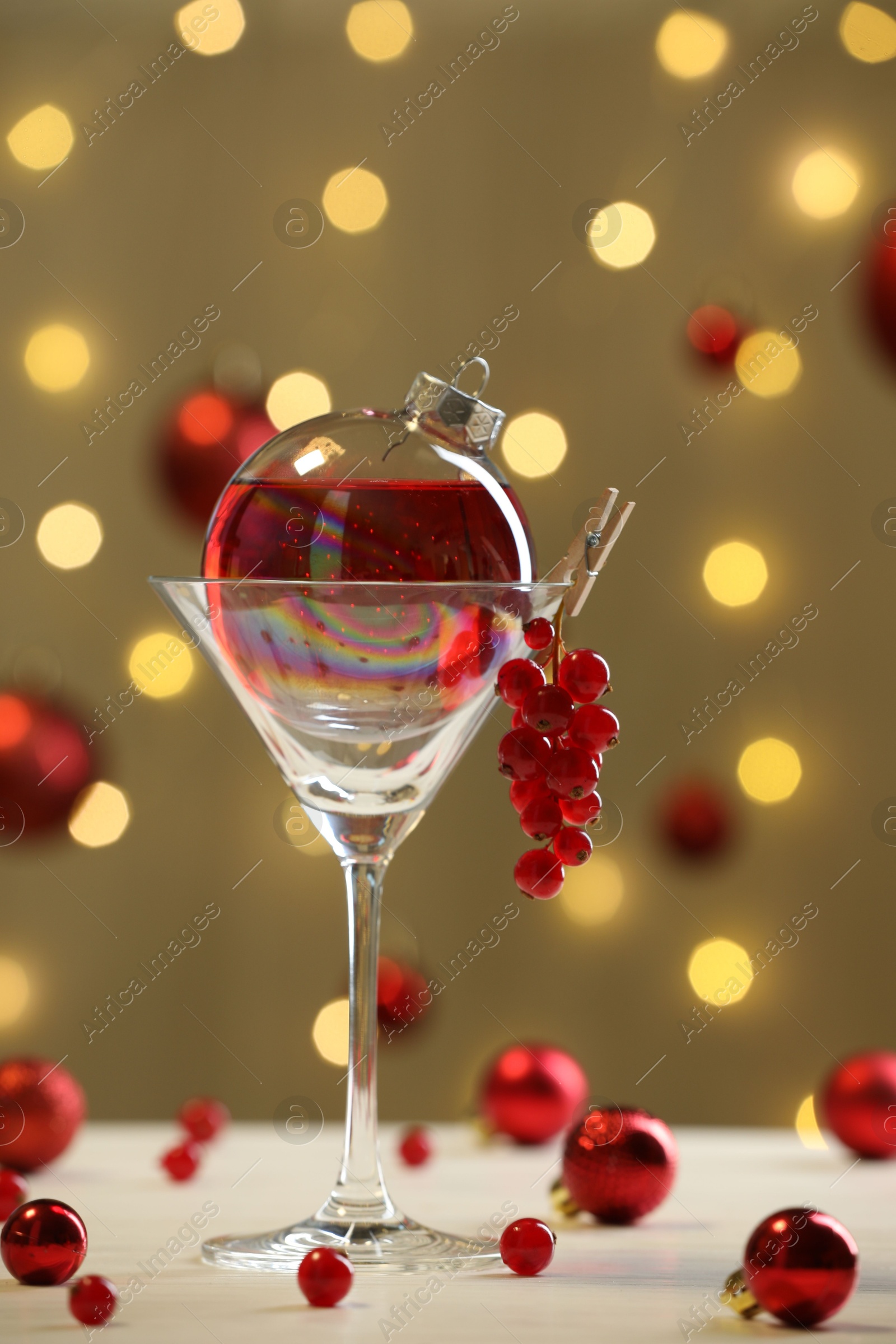 Photo of Creative presentation of Christmas cocktail in bauble and glass on white table against dark beige background with blurred lights