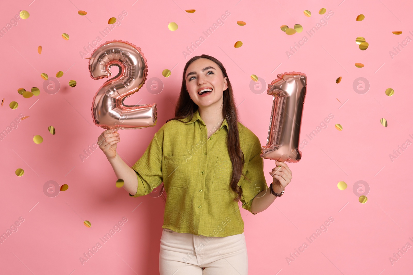 Photo of Coming of age party - 21st birthday. Happy young woman with number shaped balloons and confetti on pink background