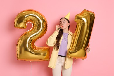 Photo of Coming of age party - 21st birthday. Happy young woman with number shaped balloons, hat and blower on pink background