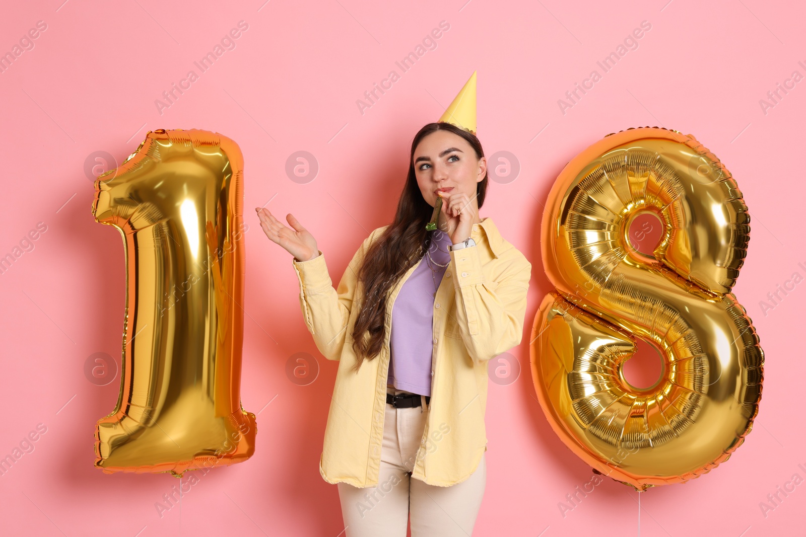 Photo of Coming of age party - 18th birthday. Happy young woman with number shaped balloons, hat and blower on pink background