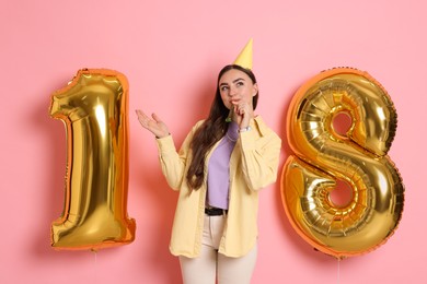 Photo of Coming of age party - 18th birthday. Happy young woman with number shaped balloons, hat and blower on pink background