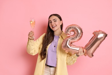 Photo of Coming of age party - 21st birthday. Happy young woman with number shaped balloons and glass of sparkling wine on pink background