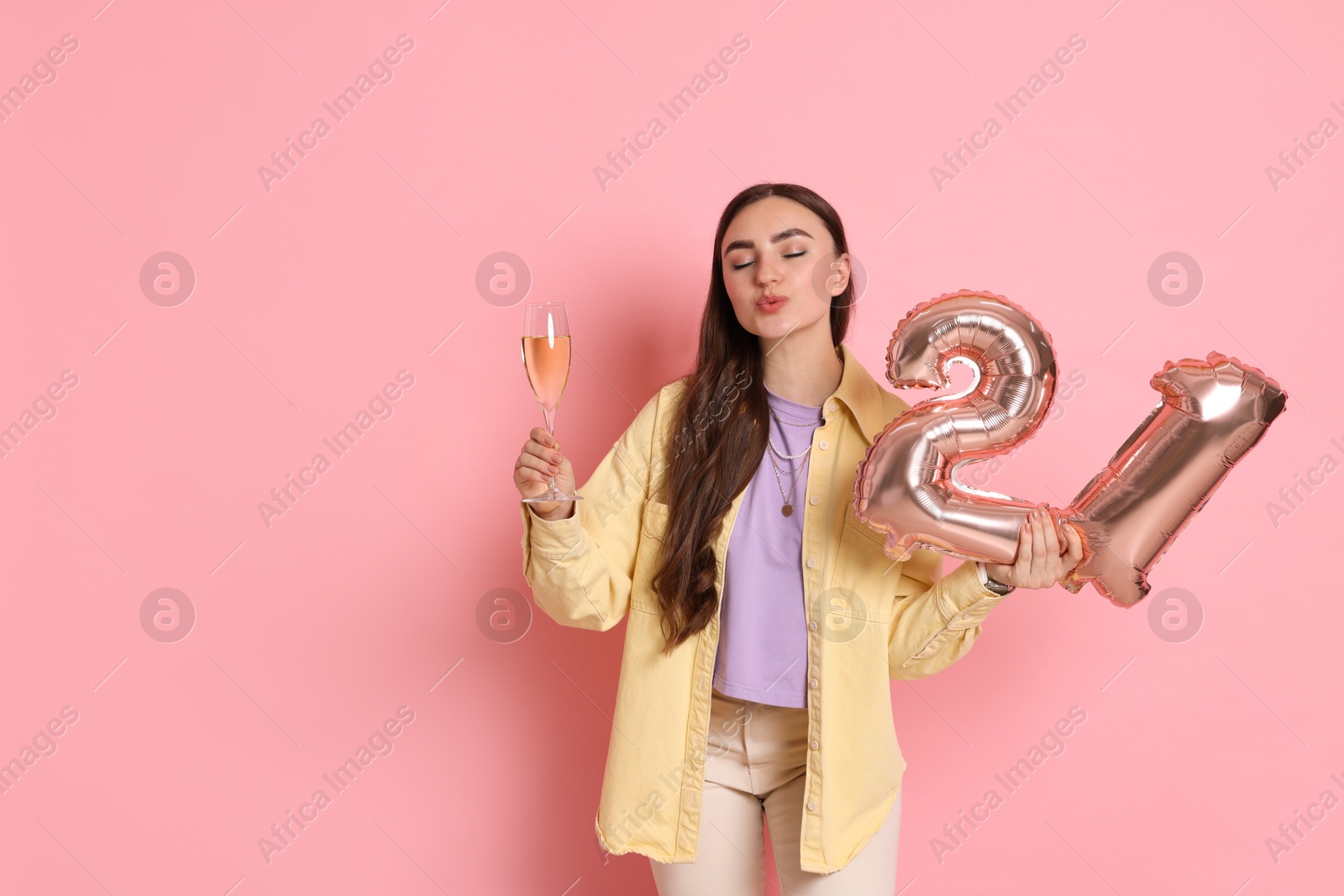 Photo of Coming of age party - 21st birthday. Happy young woman with number shaped balloons and glass of sparkling wine on pink background, space for text